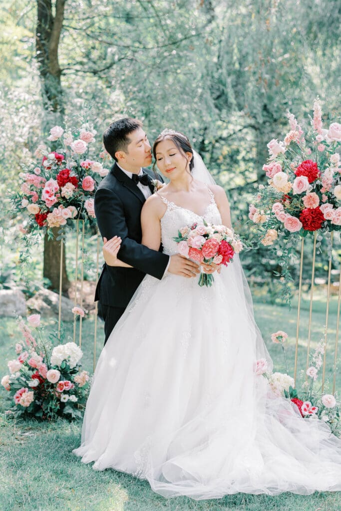 A bride and groom standing outdoors, surrounded by the enchanting tones of Valentine’s-inspired floral arrangements in shades of red, pink, and cream.

